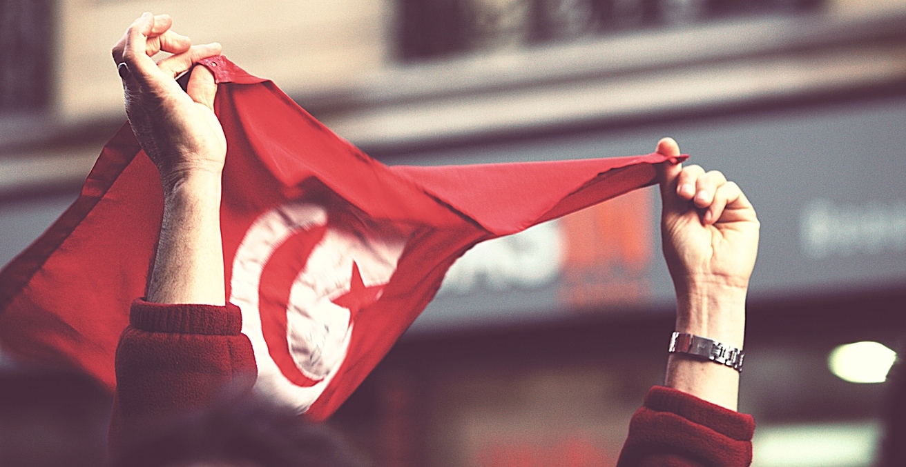 A Tunisian flag flies at a protest in 2011. Source: Gwenael Piaser, Flickr, https://bit.ly/3HWhmHN.